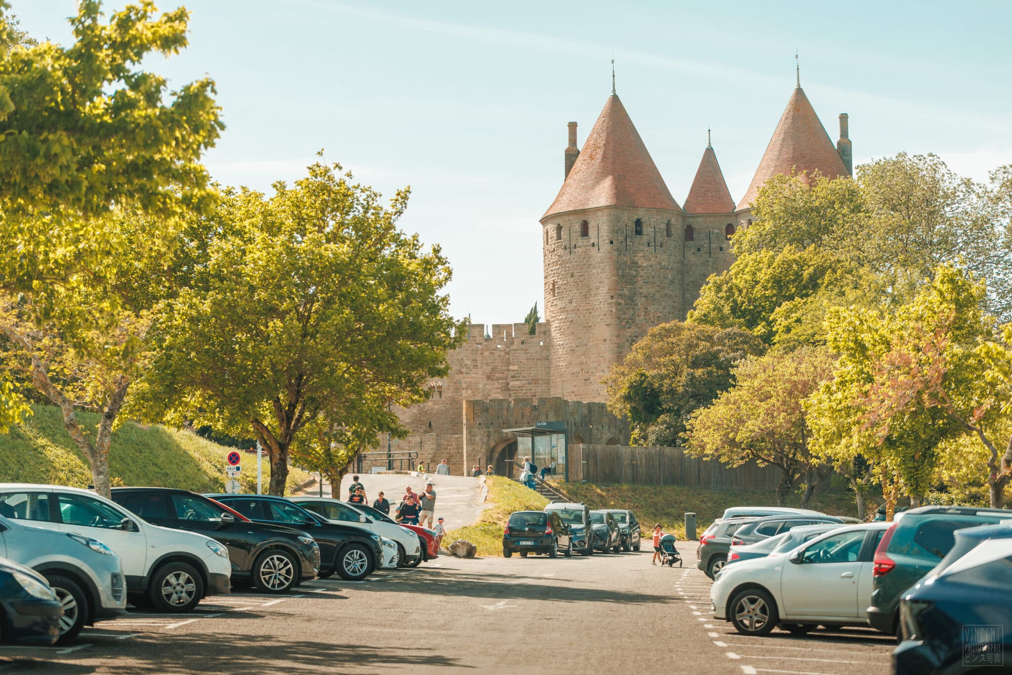des voitures sont au premier plan puis la porte narbonnaise de la cité médiévale se dresse derrière les arbres